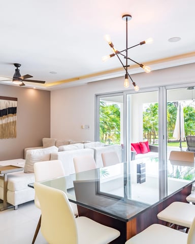 Modern open-plan living and dining area with cream sofa, dining table, glass doors opening to patio with palm trees and lounge chairs